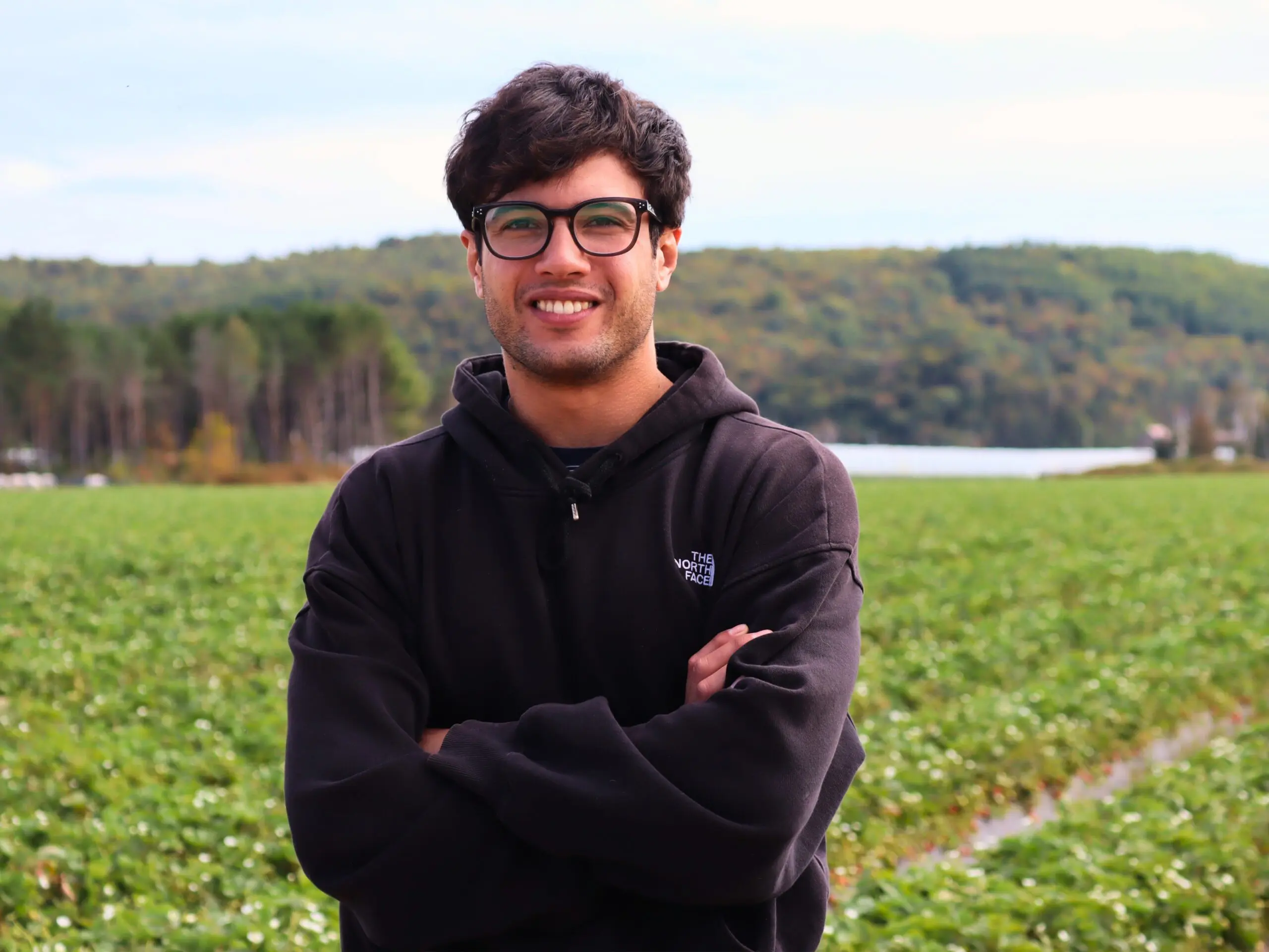 Un jeune homme aux cheveux foncés, portant des lunettes et un sweat à capuche noir, se tient debout, les bras croisés, et sourit devant un champ vert et des arbres, sous un ciel nuageux.