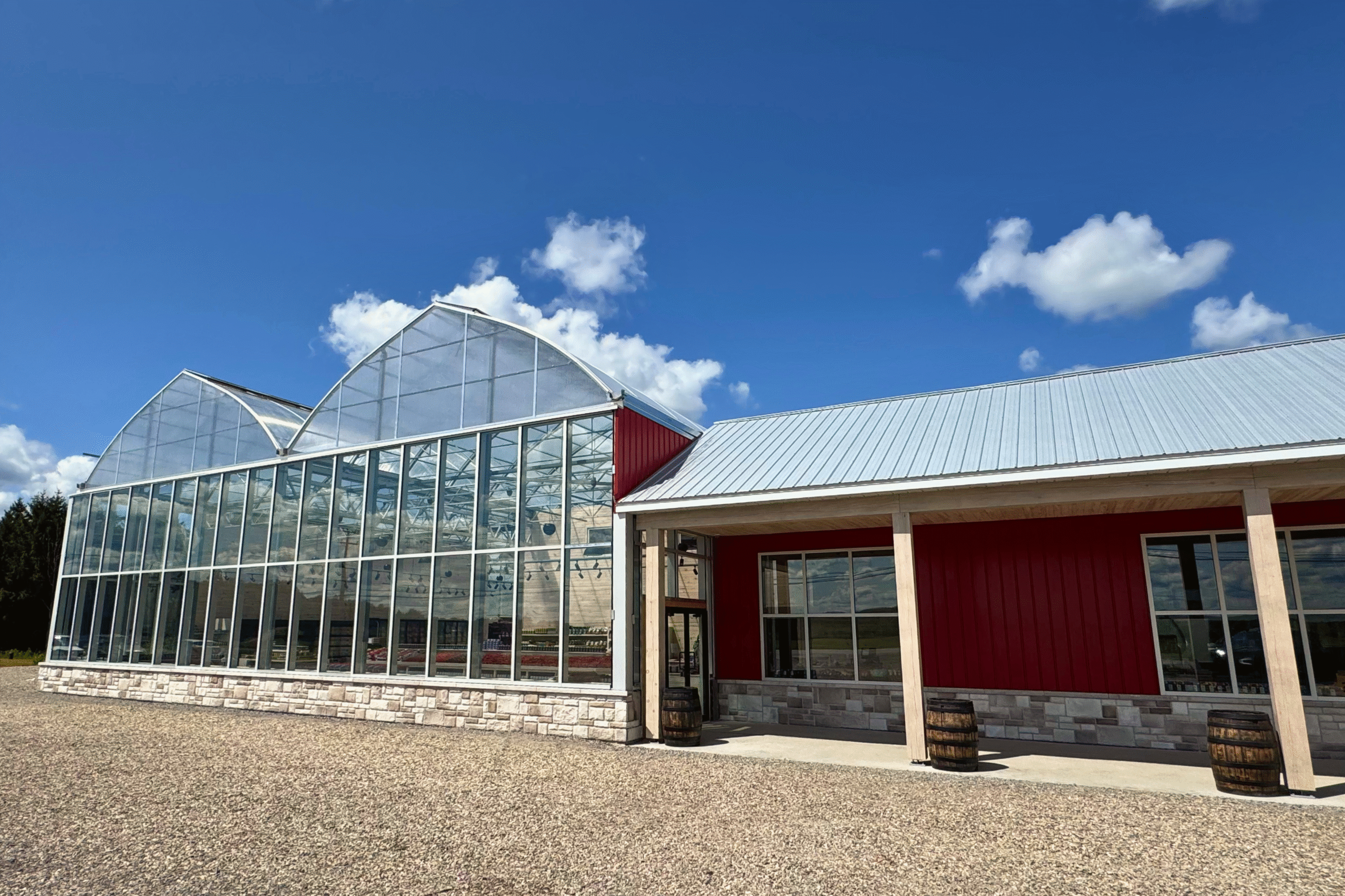 Un bâtiment moderne avec une grande serre en verre et une structure rouge et blanche avec un toit en métal, le tout sous un ciel bleu avec des nuages épars. Deux tonneaux en bois se trouvent près de l'entrée.