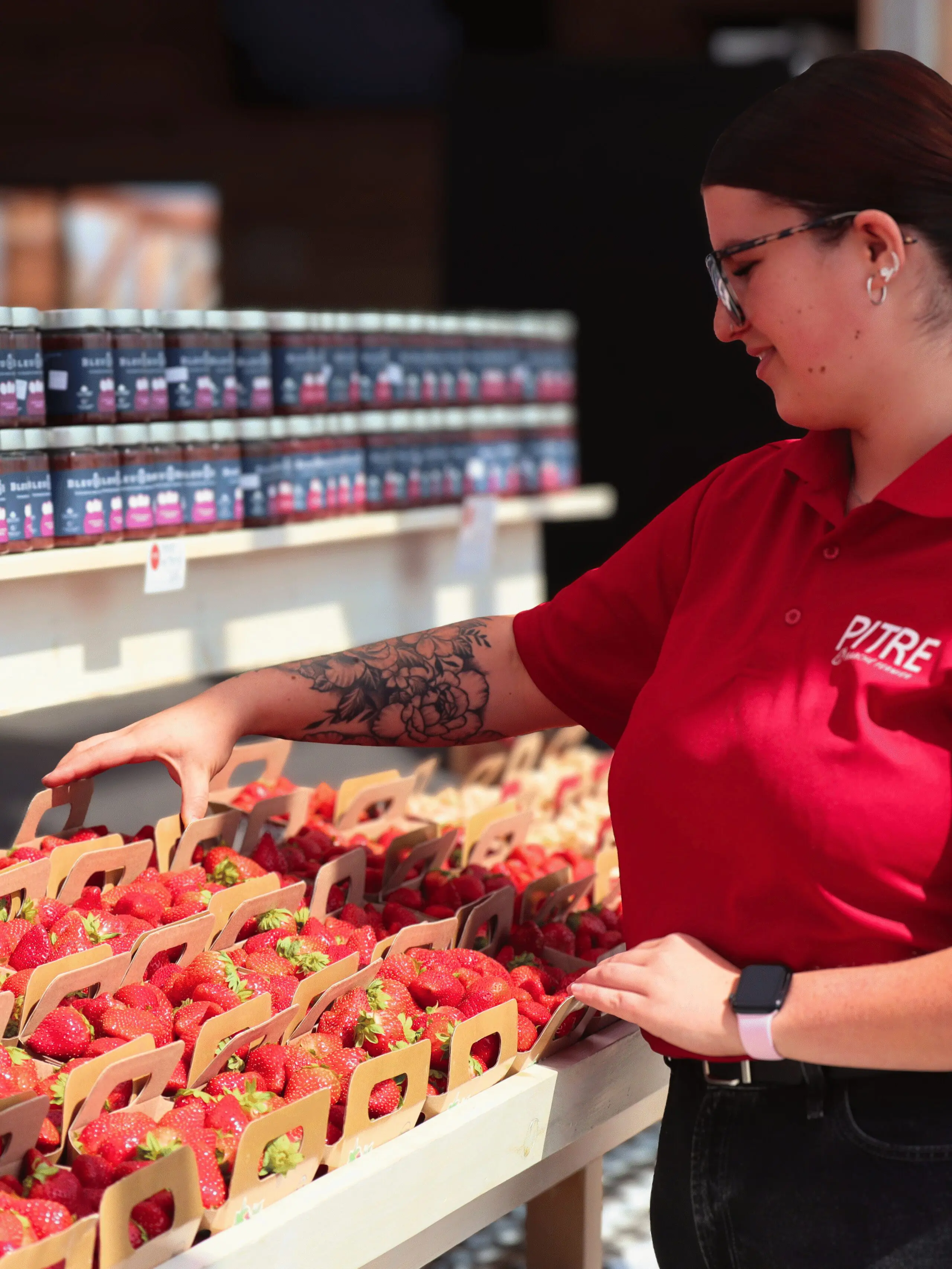 Une personne en chemise rouge au bras tatoué dispose des paniers de fraises fraîches sur un présentoir dans un marché, avec des bocaux de conserves alignés sur des étagères à l'arrière-plan.