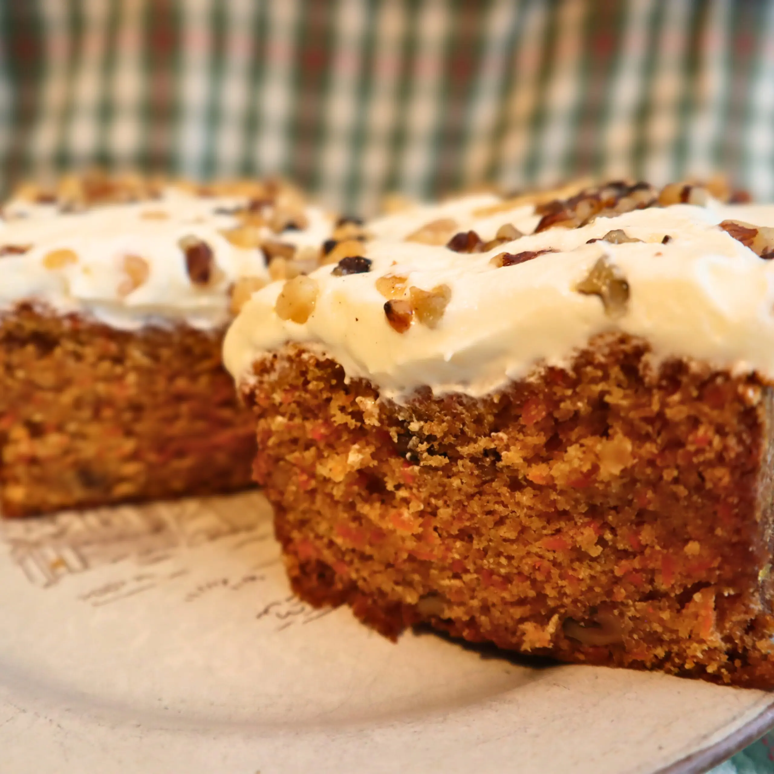 Close-up of two thick slices of carrot cake topped with creamy white frosting and chopped nuts, placed on a patterned plate with a blurred plaid fabric in the background.