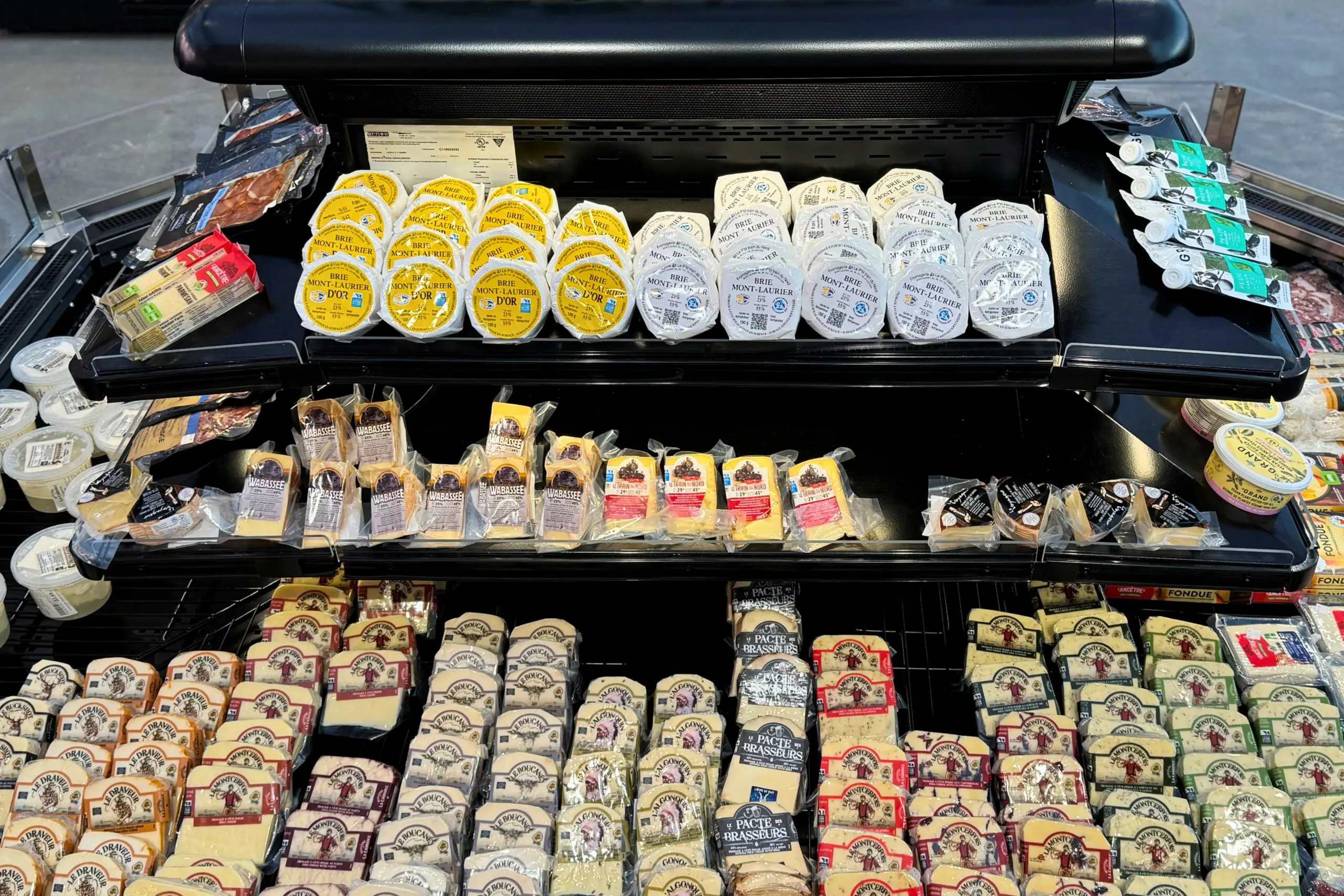 A refrigerated grocery display filled with various types of packaged cheeses, including round containers and rectangular blocks, arranged neatly on black shelves.