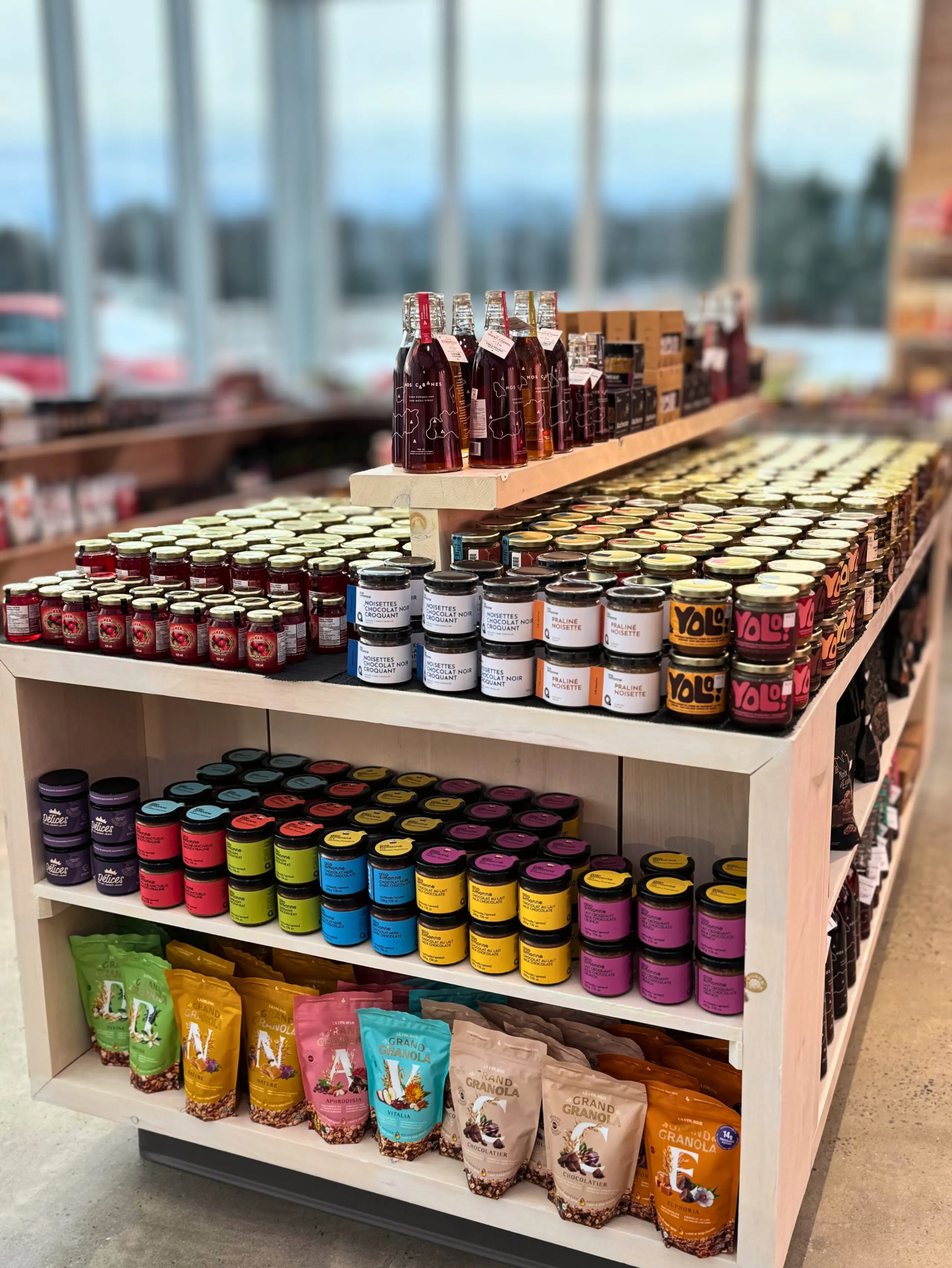 A display table in a store with neatly arranged jars of preserves, colorful packaged snacks, and bottles of drinks, set against large windows showing an outdoor view.