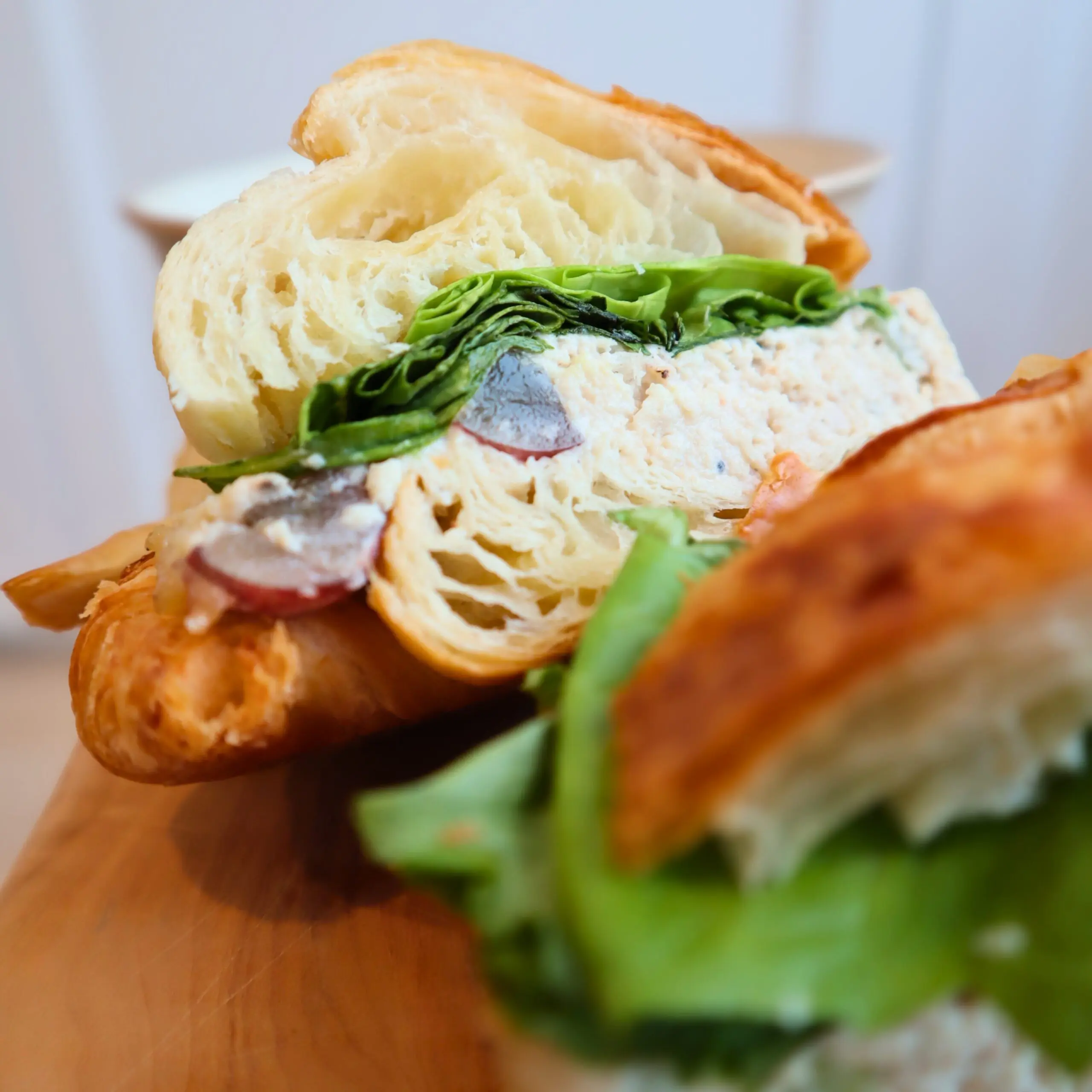 A close-up of a croissant sandwich filled with leafy greens, sliced grape tomatoes, and a creamy chicken salad on a wooden surface. The croissant appears fresh and flaky.