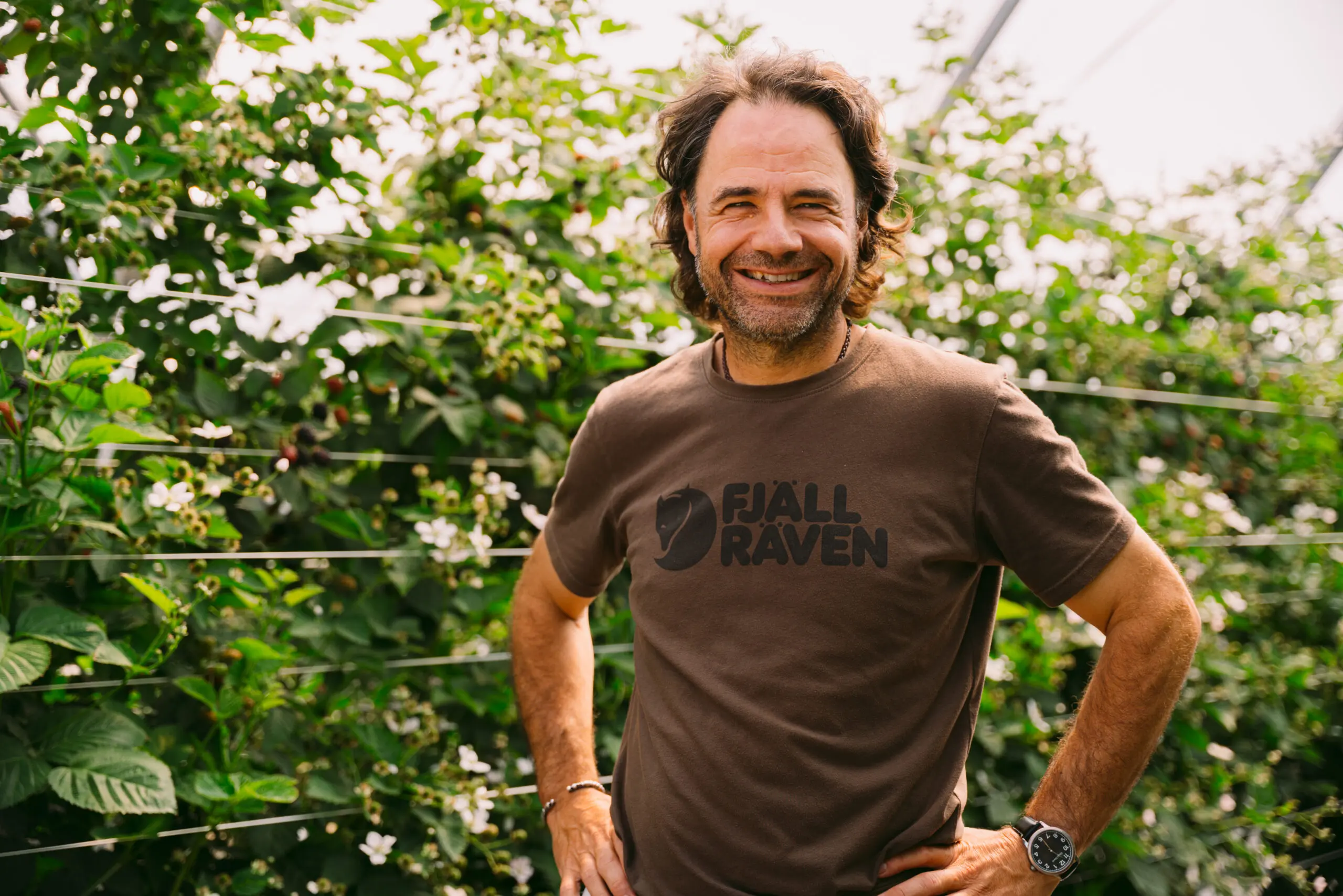 Un homme aux cheveux longs et à la barbe sourit alors qu'il se tient à l'extérieur parmi des plantes vertes, vêtu d'un t-shirt Fjällräven marron et d'une montre. La lumière du soleil filtre à travers le feuillage en arrière-plan.
