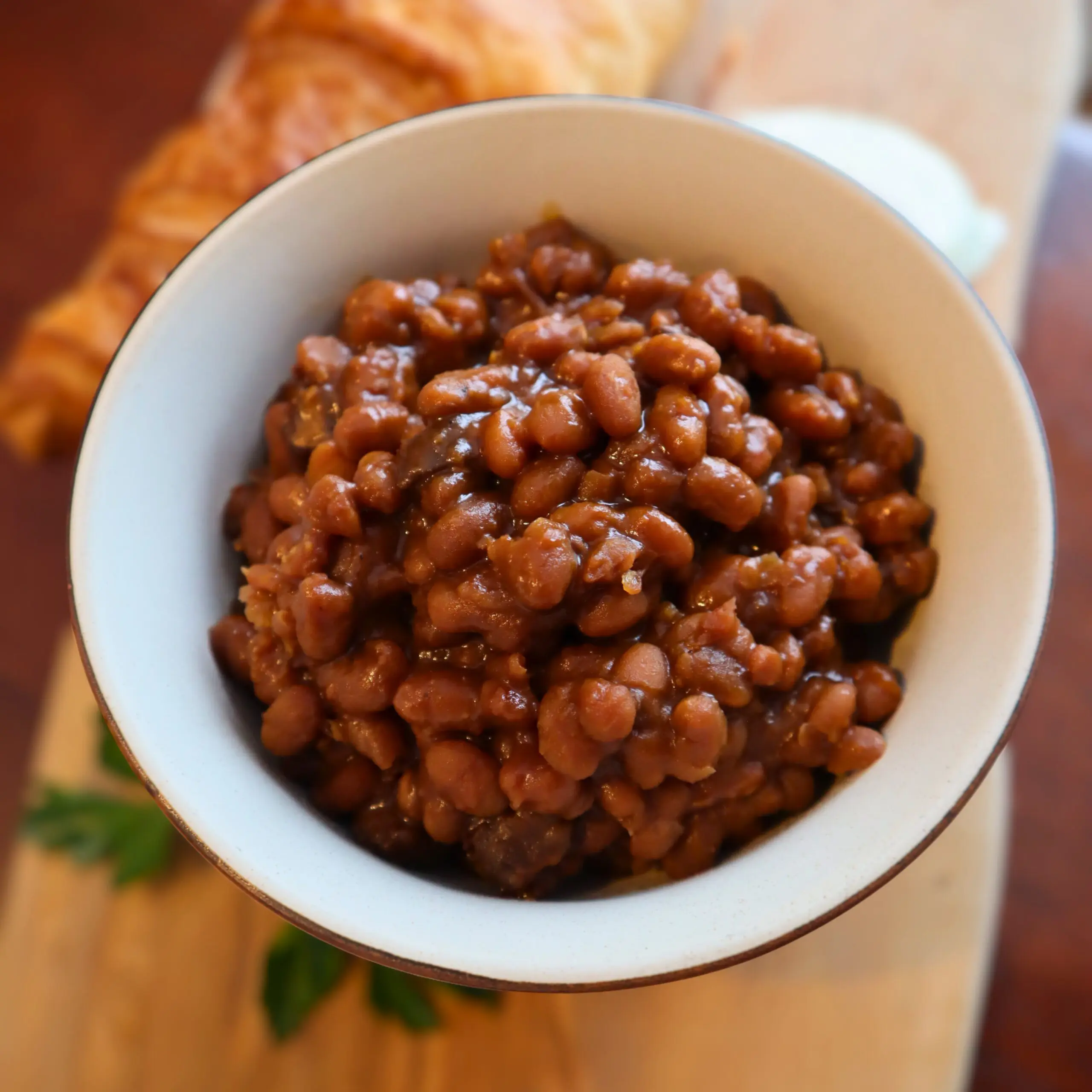 A white bowl filled with baked beans in a rich brown sauce sits on a wooden surface, with a croissant and garnish blurred in the background.