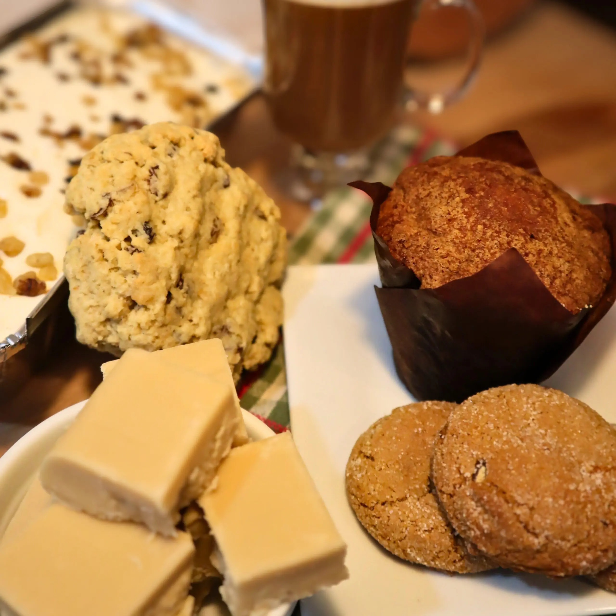 A plate with vanilla fudge squares, a large muffin, cookies, a chunk of cookie dough, and a tray of dessert in the background, with a glass of coffee in the back.