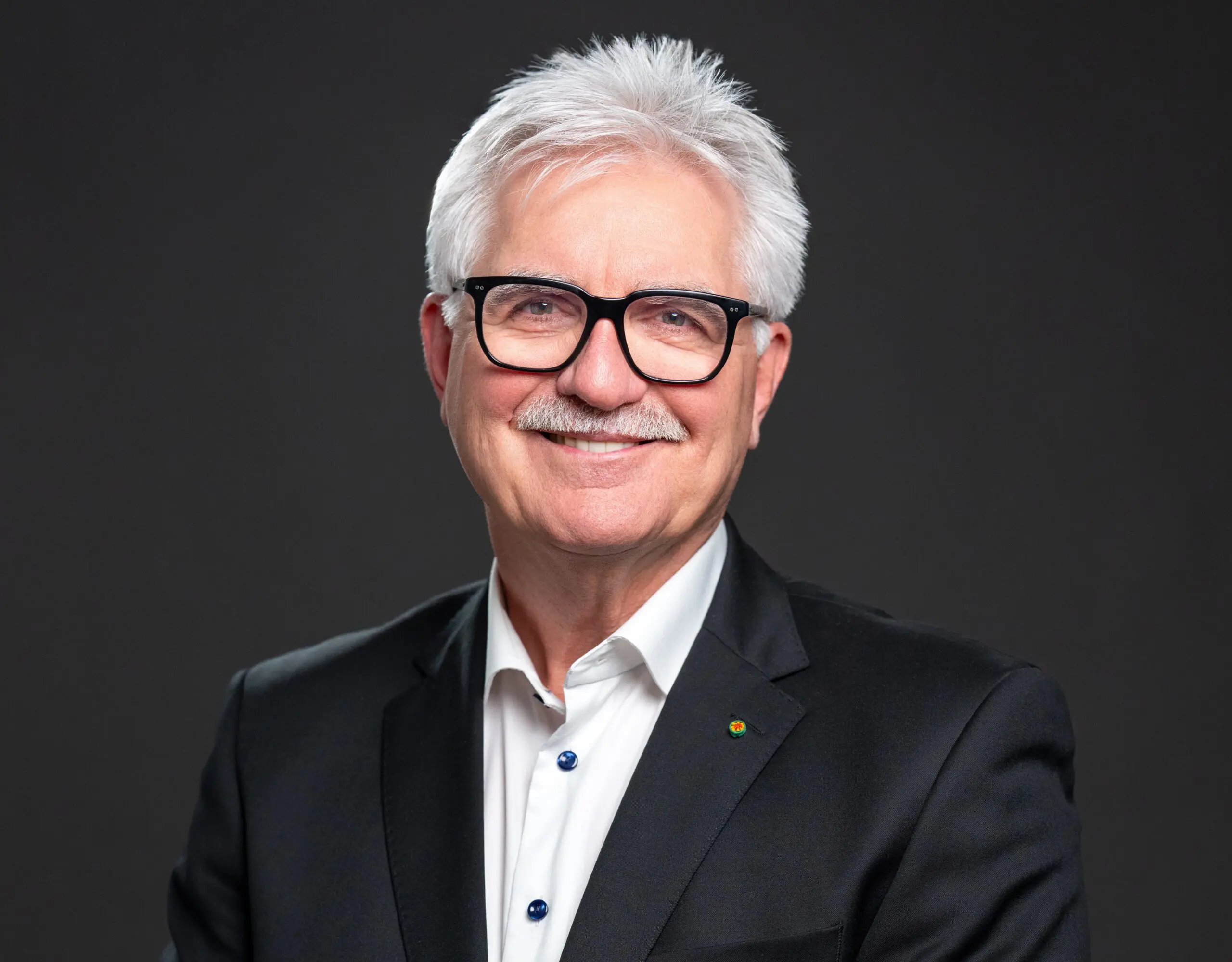 A smiling older man with white hair, glasses, and a mustache, wearing a black suit jacket and white shirt, poses against a plain dark background.