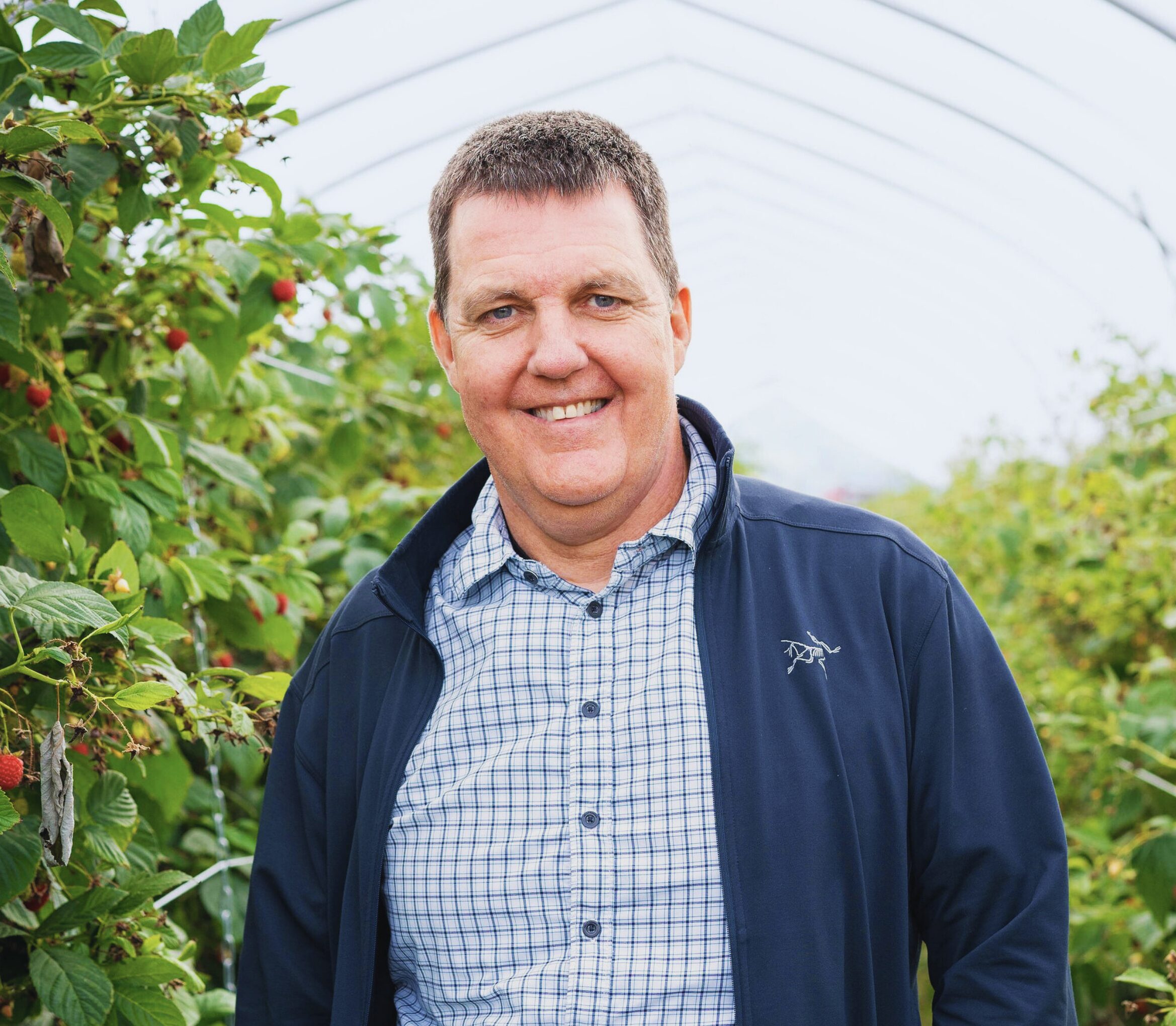 A man in a blue jacket and checkered shirt stands smiling in a greenhouse surrounded by green plants with red berries. The greenhouse structure arches overhead, and the scene appears bright and airy.