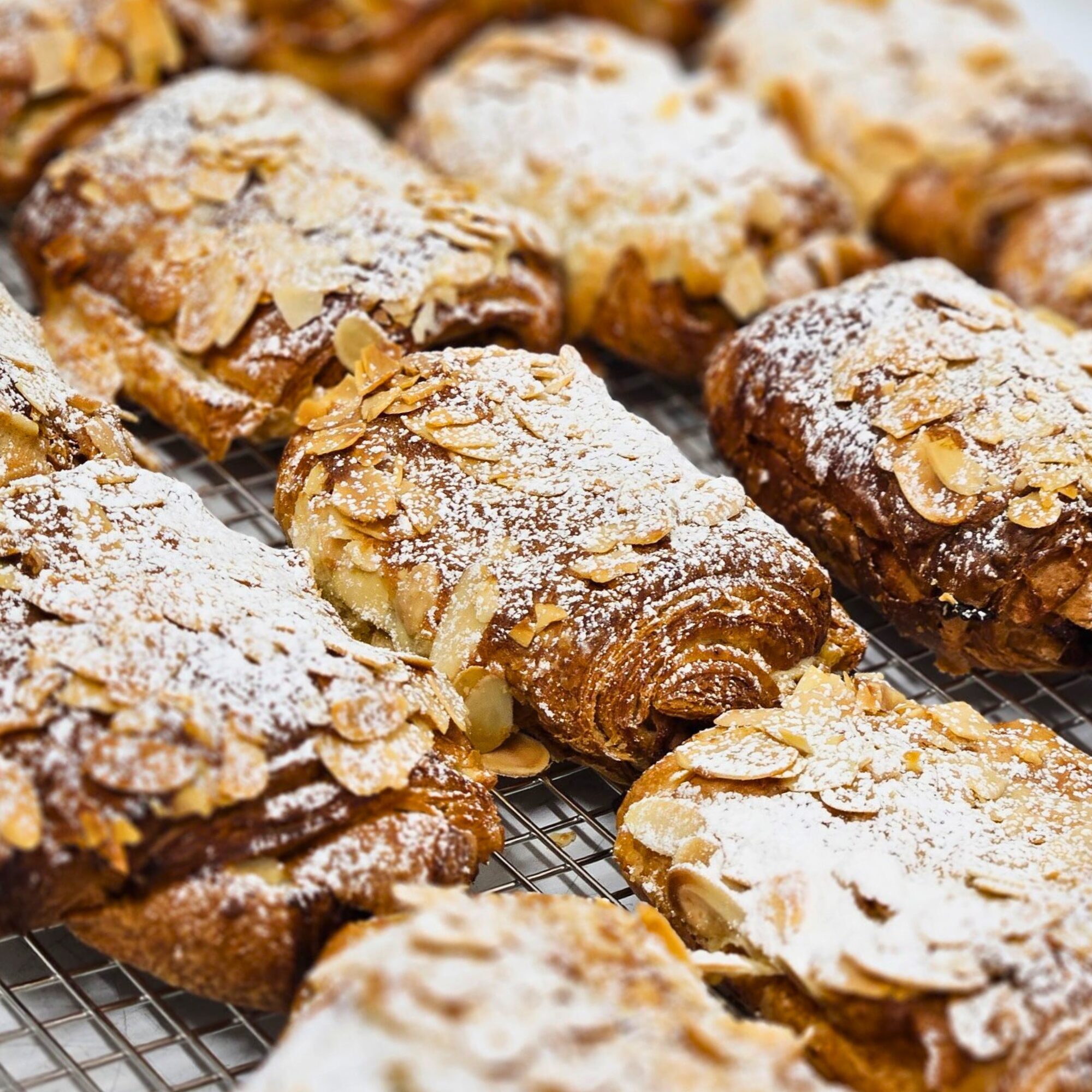 Des rangées de pâtisseries feuilletées garnies d'amandes tranchées et de sucre en poudre refroidissent sur une grille. Les pâtisseries dorées ont une texture superposée et croustillante et semblent fraîchement cuites.