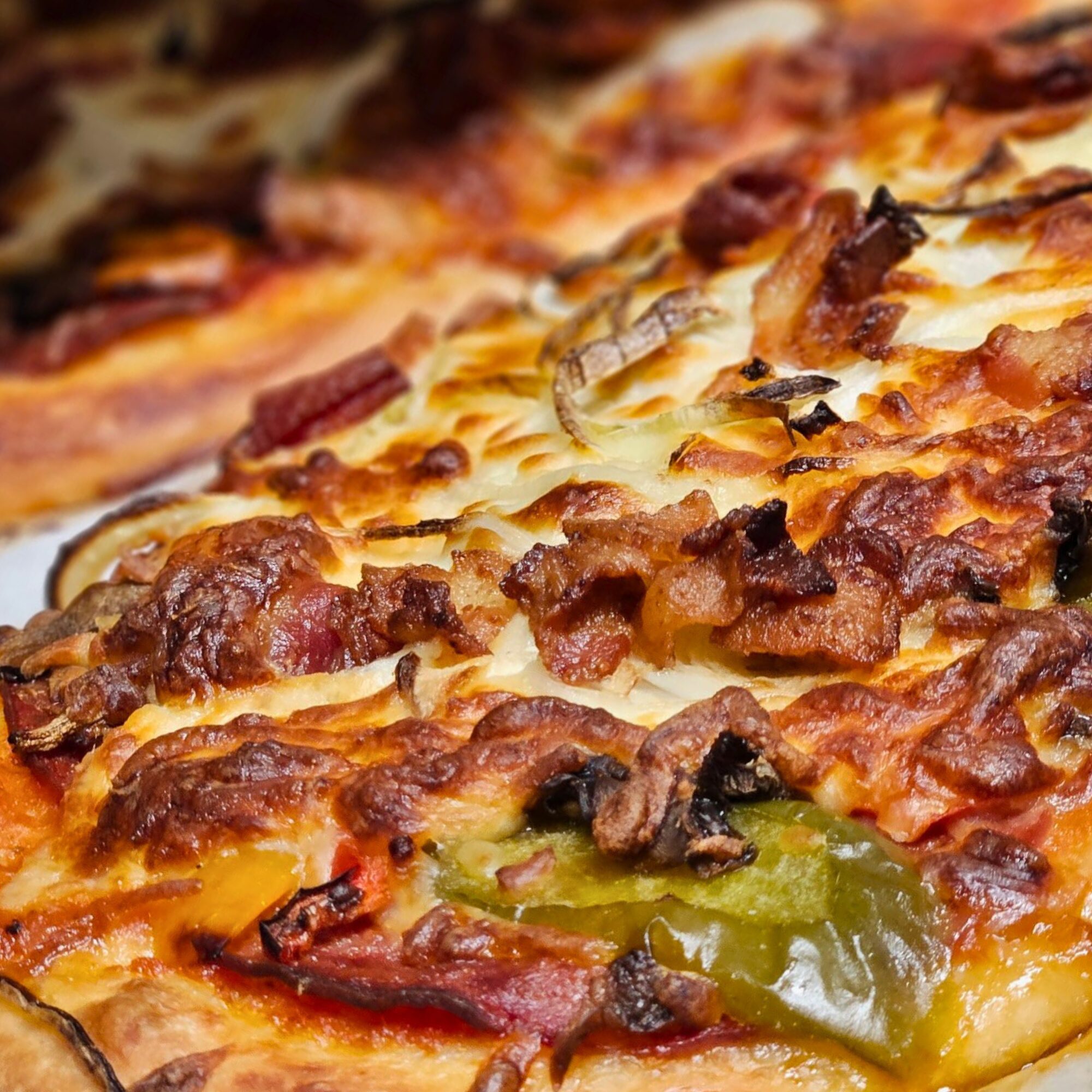 Close-up of a baked pizza topped with melted cheese, pieces of bacon, sliced green bell peppers, onions, and tomato sauce. The crust appears golden and crispy.