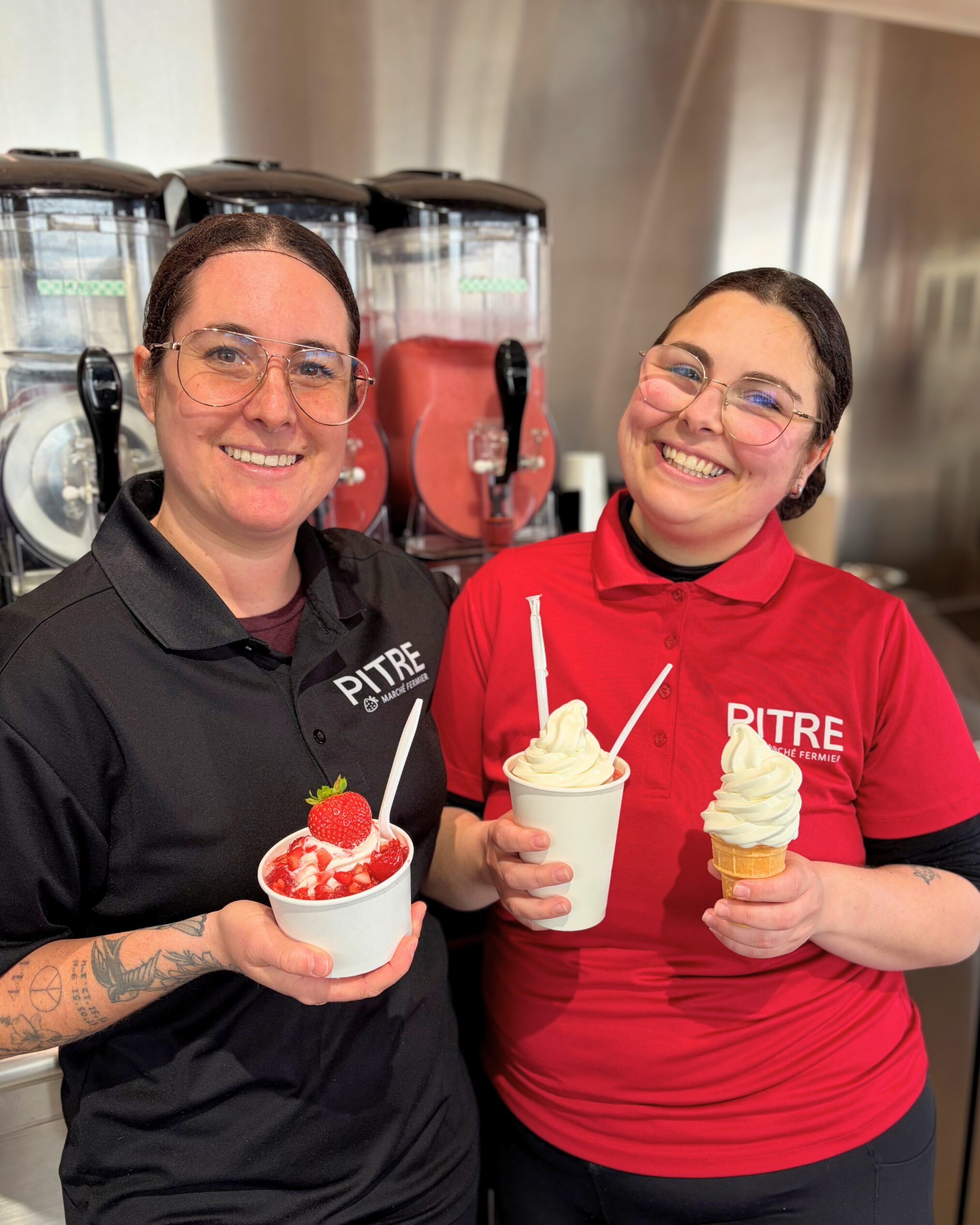 Deux femmes souriantes portant des chemises de la marque Pitre se tiennent devant des machines à slush. L'une tient une couple glacée aux fraises, l'autre un Mont-Blanc et un cornet de crème glacée molle.